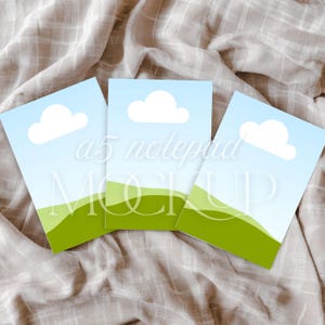 May include: Three rectangular notepads featuring a graphic illustration of a light blue sky with a single white cloud above rolling green hills. The text "a5 notepad MOCKUP" is overlaid in white across the center of the three notepads.
