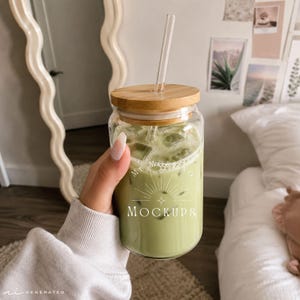 Puede incluir: Un vaso de cristal transparente con tapa de bambú y pajita de cristal recta, lleno de una bebida helada de color verde claro. El vaso presenta un diseño gráfico blanco con rayos de sol y el texto "MY MOCKUPS" impreso en la parte frontal.