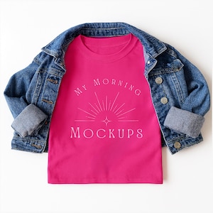 May include: A bright pink t-shirt featuring a white graphic of a sunburst and a four-pointed star with the text "MY MORNING MOCKUPS" layered underneath a blue denim jacket with silver buttons and folded cuffs, displayed on a plain white background.