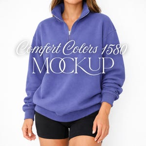 May include: A periwinkle blue sweatshirt with a quarter-zip collar and the text "Comfort Colors 1580 MOCKUP". The model is wearing black shorts. The sweatshirt has long sleeves.