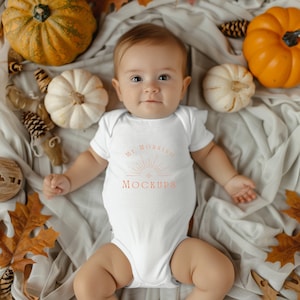 May include: A white baby onesie with the text "My Morning Mockups" in orange. The onesie is worn by a baby lying on a white blanket with fall-themed decorations.