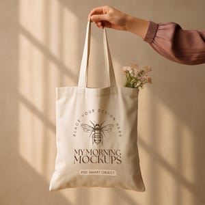 May include: A beige tote bag with a bee graphic and the text "PLACE YOUR DESIGN HERE" and "MY MORNING MOCKUPS". The bag is held by a person and contains a small bouquet of flowers.