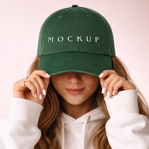 以下が含まれることがあります： 白いブロック体の「MOCKUP」という文字が入った、濃い緑色の野球帽。白いパーカーを着た人が帽子を持っています。背景はソフトピンクです。