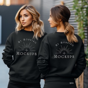 以下が含まれることがあります： 2人の女性が、前面と背面に「My Morning Mockups」と書かれた白いグラフィックデザインが施された黒いスウェットシャツを着ています。グラフィックデザインは、光線と星を持つスタイリッシュな太陽を特徴としています。