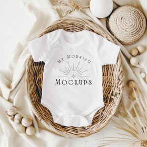 May include: A white baby bodysuit with the text "MY MORNING MOCKUPS" in black script. The bodysuit is presented in a woven basket, with decorative elements including a white sphere, wooden beads, and dried plants. The bodysuit has short sleeves and a snap closure.