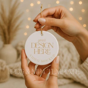May include: A round white ceramic ornament featuring the gold text YOUR DESIGN HERE and VIDEO MOCKUP, suspended by a thin gold string. The ornament has a smooth finish and a circular hole at the top for hanging, presented as a digital template.