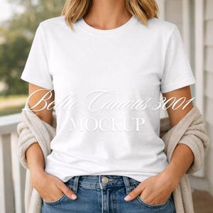 Può includere: Una maglietta bianca Bella Canvas 3001. La maglietta a maniche corte è indossata con jeans blu e un cardigan beige. Il testo "Bella Canvas 3001 MOCKUP" è visualizzato sulla parte anteriore della maglietta in corsivo.