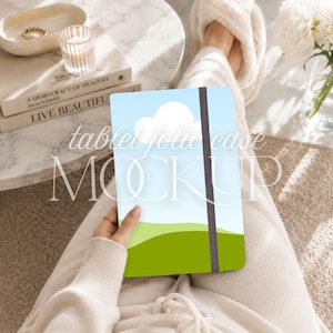 May include: A tablet folio case featuring a graphic print of a light blue sky with white clouds above a green rolling hill landscape. The case has a vertical dark grey elastic strap closure and is held in a hand, displaying the text tablet folio case MOCKUP.