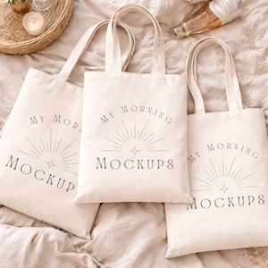 Puede incluir: Tres bolsas de tela de color blanco roto con un diseño impreso en gris oscuro que muestra un gráfico de rayos de sol y el texto MY MORNING MOCKUPS. Cada bolsa tiene dos asas de tela a juego y una forma rectangular.