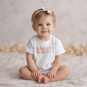 May include: A white short-sleeved t-shirt featuring the text RABBIT SKINS 3322 in a light brown, rounded font on the chest. The garment is worn with a matching light beige headband adorned with a small bow detail on the top of the head.