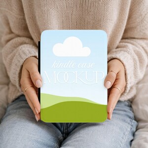 Puede incluir: Una funda rectangular para lector electrónico con un cielo azul claro y una nube blanca sobre colinas verdes. El texto kindle case MOCKUP está impreso en blanco en el centro del diseño. La funda tiene esquinas redondeadas.
