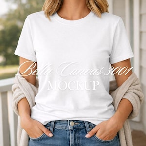 Puede incluir: Una camiseta blanca de cuello redondo con el texto "Bella Canvas 3001 MOCKUP" en cursiva. La modelo lleva vaqueros azules y una rebeca beige. Las manos están en los bolsillos de los vaqueros.