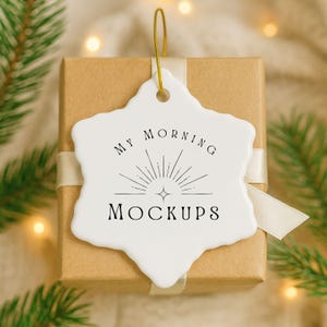 May include: A white ceramic ornament with the text "MY MORNING MOCKUPS" in black, hanging by a gold string. The ornament is snowflake-shaped and rests on a brown gift box tied with a cream ribbon.