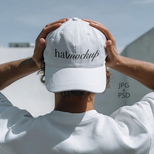 Op de afbeelding: Een witte baseballcap met de tekst "hatmockup" in zwart schrift. De pet wordt door een persoon omhoog gehouden. De achtergrond is een lichtblauwe lucht en een witte muur.