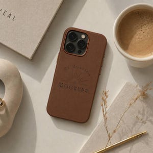 以下が含まれることがあります： 背面に太陽の光のグラフィックと「MY MORNING MOCKUPS」の文字が刻印された茶色のスマートフォンケース。ケースにはトリプルレンズカメラシステム用の切り欠きがあり、コーヒーカップの近くの明るい色の表面に置かれています。