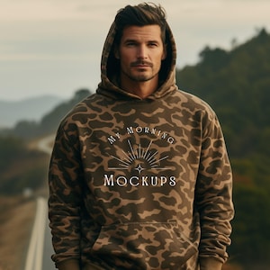 Puede incluir: Una sudadera con capucha de camuflaje marrón con capucha con cordón. La sudadera con capucha muestra el texto "MY MORNING MOCKUPS" en blanco. El fondo es una escena borrosa al aire libre con una carretera y árboles.