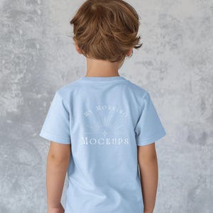 May include: A pale blue t-shirt with the text "MY MORNING MOCKUPS" in white, worn by a child. The text is centered on the back of the shirt, above a sunburst design. The child is facing away from the camera, with brown hair.