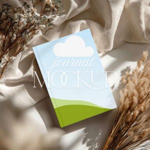 May include: A journal mockup with a light blue cover featuring a white cloud and green landscape design. The words "journal MOCKUP" are printed on the cover. The journal is placed on a cream-colored fabric with dried plants.