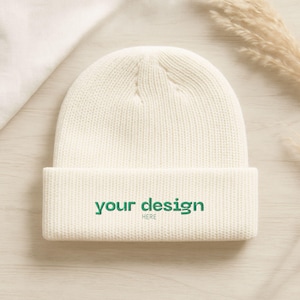 May include: Cream-colored beanie with a folded brim. The text "your design HERE" is printed in green on the front. The hat is made of a knitted material and is displayed on a light wooden surface.