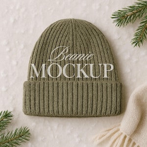 May include: Olive green ribbed knit beanie with the text "Beanie MOCKUP" in white script. The hat is displayed on a textured white surface, with a sprig of evergreen and a cream-colored scarf in the background.