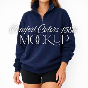 Puede incluir: Una maqueta de sudadera azul marino con cremallera de un cuarto con el texto "Comfort Colors 1580 MOCKUP" en blanco. La sudadera tiene un cuello alto y una cremallera plateada. La modelo lleva unos pantalones cortos negros.