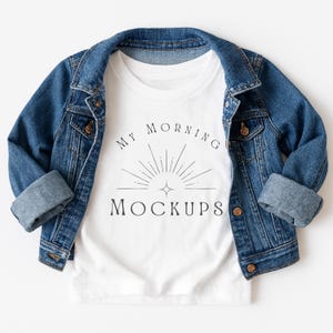 Può includere: Una maglietta bianca con la scritta "MY MORNING MOCKUPS" in nero, sovrapposta a una giacca di jeans blu. La giacca ha un design classico con bottoni e maniche arrotolate. La maglietta è centrata e la giacca è drappeggiata sopra.