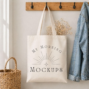 May include: A cream-colored canvas tote bag featuring black text that reads MY MORNING MOCKUPS above a graphic of a sunburst with a four-pointed star. The bag has two long shoulder straps and is hanging from a wooden hook on a wall.