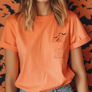 May include: An orange short-sleeved t-shirt with a chest pocket. The pocket features a black graphic of flying bats above the text MY MORNING MOCKUPS with a sunburst design. The sleeves are cuffed and the fabric has a smooth, solid texture.