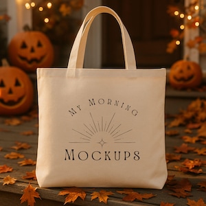 May include: Beige canvas tote bag with the text "MY MORNING MOCKUPS" in black. The bag has two handles and is sitting on a wooden surface with fall leaves and carved pumpkins in the background. The scene evokes a festive autumn theme.
