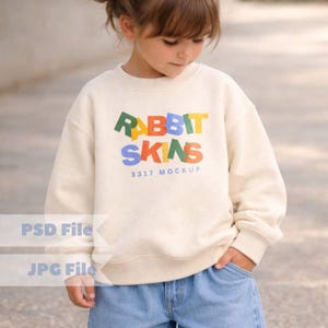 Puede incluir: Una sudadera color crema con el texto "RABBIT SKINS" en letras de colores. Debajo del texto está "3317 MOCKUP". La sudadera la lleva un niño con vaqueros azules.