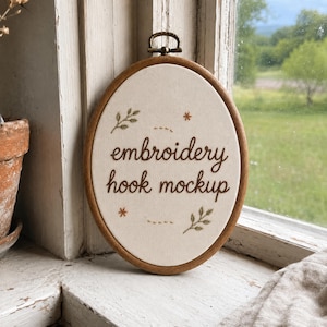 May include: An oval wooden embroidery hoop containing off-white fabric with the brown embroidered text "embroidery hook mockup". The design features small green leaf sprigs and orange star-shaped accents, with dashed lines above and below the text.