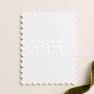 May include: A rectangular white greeting card with a scalloped edge design. The center of the card features the white text greeting card MOCKUP in a combination of script and sans-serif fonts. The card is positioned against a neutral background.