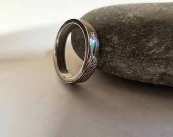 May include: A silver ring with a patterned design. The ring is sitting on a gray rock.