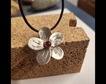 May include: Silver flower pendant necklace with a dark brown leather cord. The pendant features a flower design with five petals and a small, round, red gemstone in the center. The necklace is displayed on a brown cork surface.