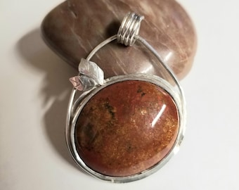 May include: A silver pendant with a red and brown jasper stone set in a silver frame. The pendant has a delicate silver leaf design.