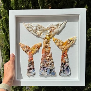 Puede incluir: Una pieza de arte de pared enmarcada en blanco con tres colas de sirena hechas de conchas marinas. Las colas están dispuestas en un patrón degradado, comenzando con conchas blancas en la parte superior y pasando a conchas azules en la parte inferior.