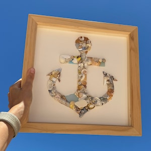 Puede incluir: Una obra de arte enmarcada que presenta un gran ancla hecha de conchas marinas. El ancla es de color marrón claro con acentos blancos y azules. El marco es de madera marrón claro.