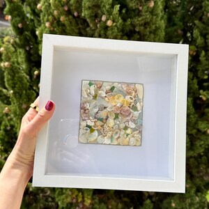 Puede incluir: Un marco blanco con una pieza de arte cuadrada en su interior. El arte está hecho de una variedad de conchas marinas en tonos de blanco, rosa y beige.