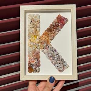 Può includere: Una lettera K incorniciata realizzata con conchiglie di vari colori e dimensioni. La lettera è esposta su uno sfondo bianco con una cornice color crema.