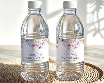 May include: Two clear plastic water bottles with white caps. Each bottle has a label with floral designs in purple, pink, and yellow, and the text "Amelia's BABY SHOWER Thanks for Coming!" The bottles sit on a woven mat.