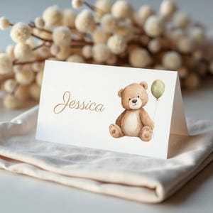 May include: A folded place card with the name "Jessica" in gold script. A watercolor illustration of a teddy bear holding a green balloon is on the right. The card is on a cream-colored cloth.