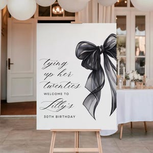 May include: A white sign on a wooden easel with a black bow illustration and text that reads "Tying up her twenties, Welcome to Lilly's, 30th Birthday." White balloons are suspended above the sign.