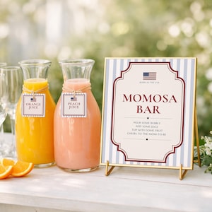 May include: Two glass carafes filled with orange and peach juice, each with a tag labelled Orange Juice or Peach Juice, next to a framed sign that reads MOMOSA BAR, BORN IN THE USA, and instructions to pour bubbly, add juice, top with fruit, and cheers.