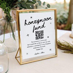 Op de afbeelding: Een goudkleurig omlijst bord met de woorden "honeymoon fund" in zwart schrift. Het bord bevat een QR-code en de tekst "Your presence is all we need..." en "Thank You". Het bord staat op een wit oppervlak.
