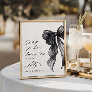 May include: A framed sign with a gold border features a black bow illustration and elegant script. The text reads "Tying up her twenties, Welcome to Lilly's 30th Birthday." The sign is on a white tablecloth, with cocktails in the background.