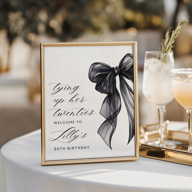May include: A framed sign with a gold border features a black bow illustration and elegant script. The text reads "Tying up her twenties, Welcome to Lilly's 30th Birthday." The sign is on a white tablecloth, with cocktails in the background.