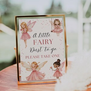 May include: A gold-framed sign with the text "a Little FAIRY Dust to go PLEASE TAKE ONE" and illustrations of fairies in pink dresses. The sign is displayed on a wooden tray.