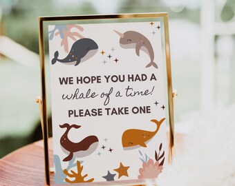 May include: A framed sign with a gold border features whimsical illustrations of whales and coral. The text reads, "WE HOPE YOU HAD A whale of a time! PLEASE TAKE ONE." The sign is set on a wooden tray, suggesting a party or event theme.