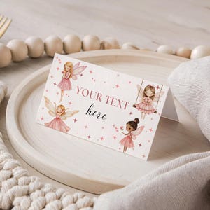 Könnte beinhalten: Eine gefaltete Tischkarte mit Aquarellillustrationen von Feen in rosa Kleidern und Flügeln. Die Karte trägt den Text "YOUR TEXT here" und ist mit rosa Sternen verziert. Die Karte liegt auf einem Holztablett.