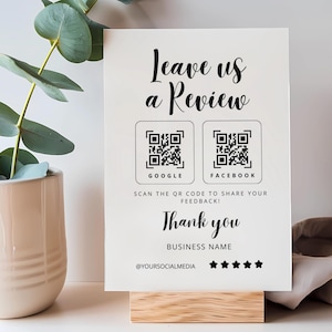 May include: White sign with black text that reads "Leave us a Review". Two QR codes are displayed below the text, one for Google and one for Facebook. The sign also includes the text "Scan the QR code to share your feedback!" and "Thank you".